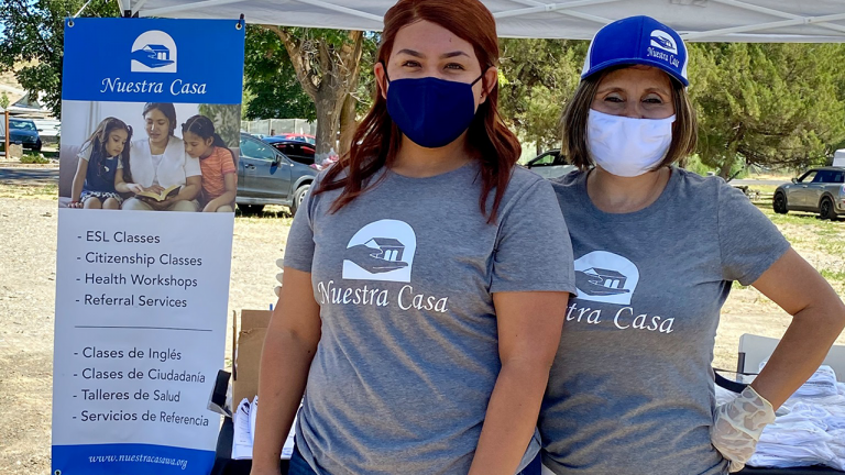 Two women in "Nuestra Casa" T-shirts and face masks stand next to a sign about resources offered by Nuestra Casa in English and Spanish (ESL Classes, Citizenship Classes, Health Workshops, Referral Services) 