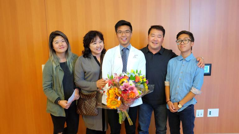 A young man with glasses and wearing a white lab coat holds bouquets of flowers and is surrounded by his family.