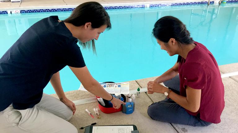 Ali, pictured on the left, assists with a swimming pool inspection.  She and another woman sit on the concrete edge of a chlorinated pool with testing equipment and a clipboard.