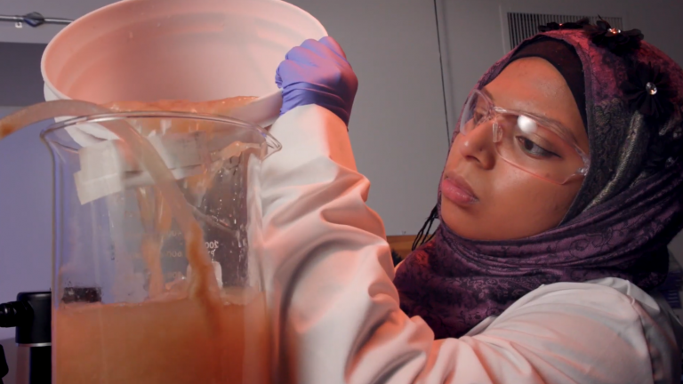 Woman in hijab and safety glasses pours tan-colored liquid from a large bucket into a large beaker.