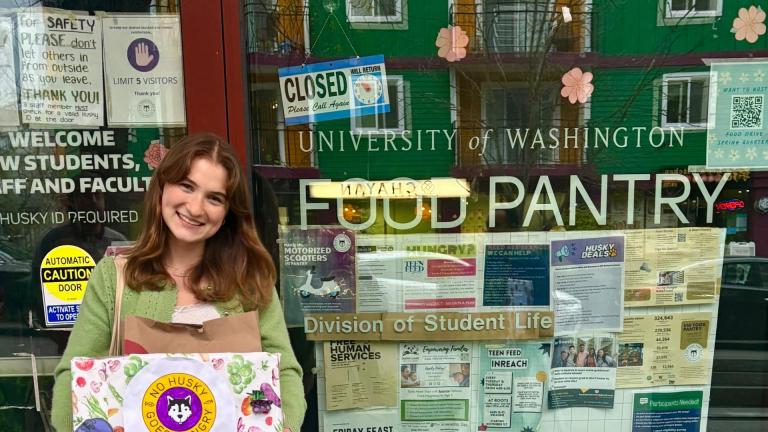 Angelina Durbin stands in front of the window of the UW Food Pantry. She is holding a cardboard box reading "No Husky Goes Hungry" with an image of a husky dog.