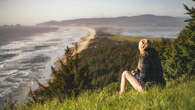 A person sits on a bluff looking out at the ocean and headlands in the distance.