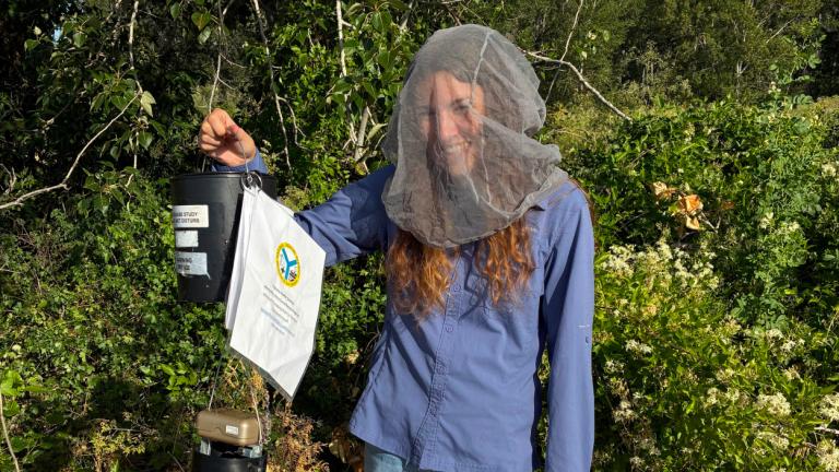 Payton Miksch holds up a mosquito trapping device while wearing a mosquito netting hood.