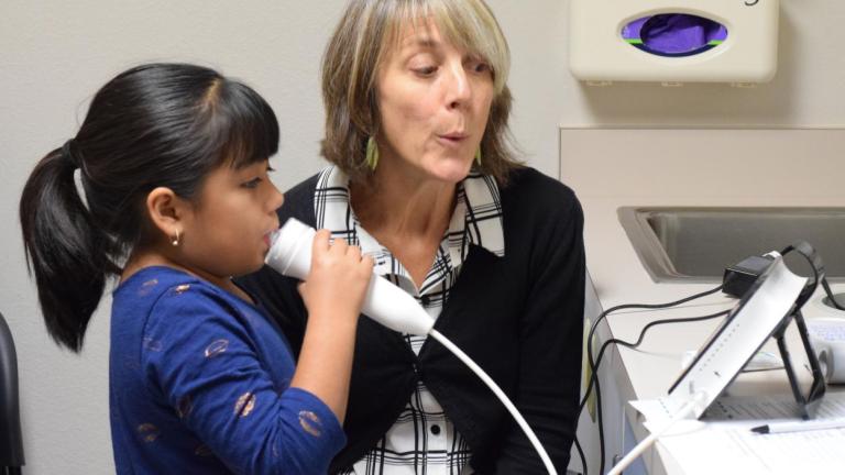 A girl blows into an asthma monitoring device while Dr. Catherine Karr demonstrates blowing sitting next to her.