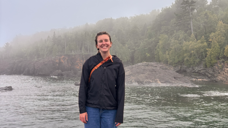 Carole Green stands in front of a lake and evergreens in fog.