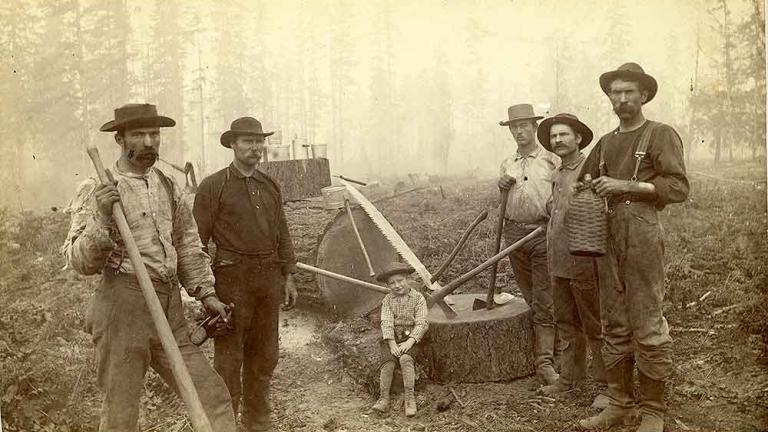 Black and white image of five loggers and a boy in Western Washington, W. T. The men are standing with axes in hand appearing to have just cut down a tree. The boy sits on the stump. 