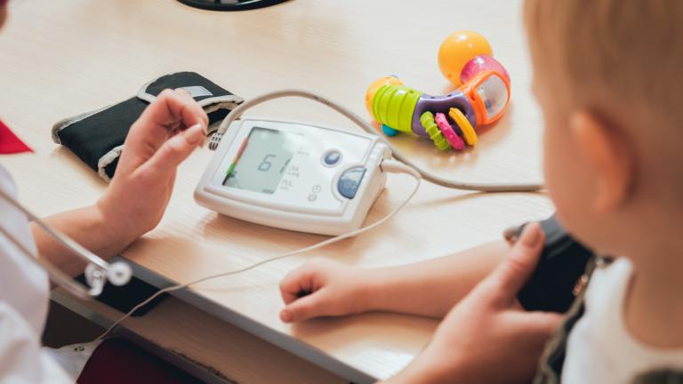 A doctor takes a child's blood pressure (only the doctor's hands and the child's arm and side of face are visible). On a table are the blood pressure monitor and a teething ring.
