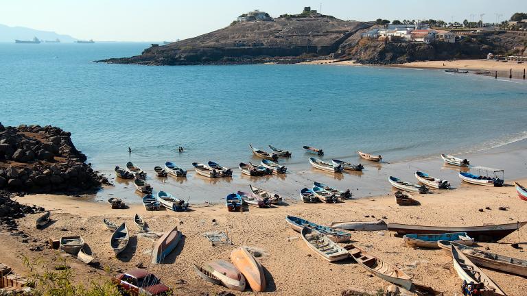 Photo of azure blue water and a beach with rowboats sitting in the sand and in the shallow water. The rocky coastline wraps around with a village up on cliffs in the distance.