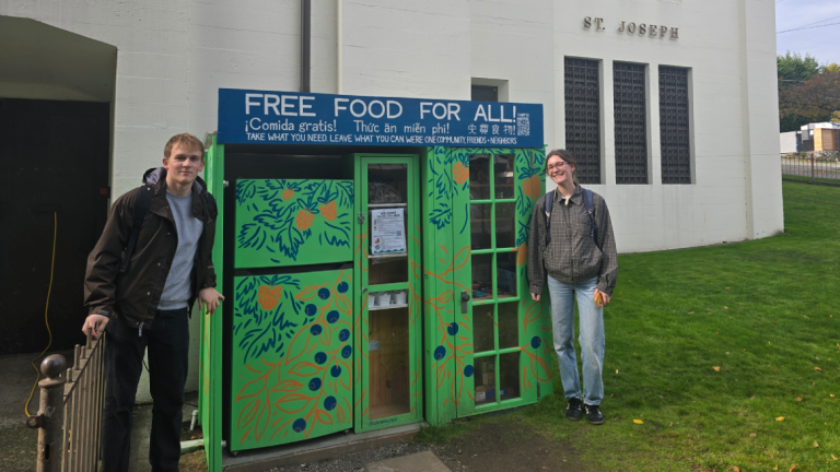 Two people stand on either side of an outdoor refrigerator and food storage cabinet. A sign above the community fridge and micropantry reads "Free food for all! (translated into Spanish, Vietnamese and Chinese) Take what you need leave what you can we're one community, friends + neighbors." The micropantry is painted green with flowers and is in front of a church labeled "St. Joseph Parish." 