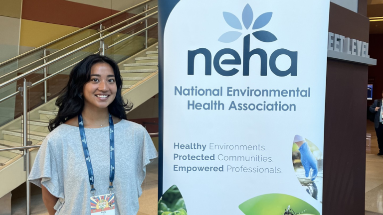 Emily Do stands in front of a large sign reading "NEHA - National Environmental Health Association. Healthy Environments. Protected Communities. Empowered Professsionals."