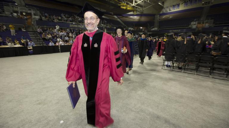 A man wearing graduation regalia leads a group of faculty out of a graduation ceremony.