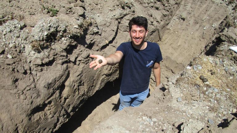 Gombart standing in a hole surrounded by rocks and soil, holding out a ball of soil in his extended right hand.