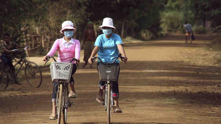 Two women wearing air masks on bicycles on a dirt road.