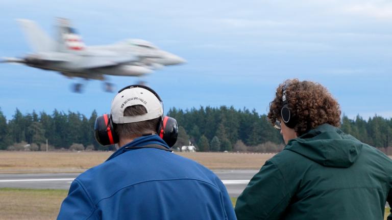 Two men facing away from the camera watch a blurred jet land on an airstrip. The men are both wearing over-ear headphones.