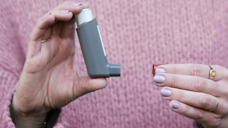 Person wearing pink sweater holds an asthma inhaler in one hand and red cap in the other.