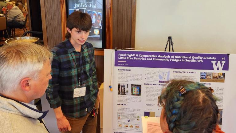 Kaidan St Louis wears a green and blue plaid shirt and stands in front of a research poster, explaining it to two observers.