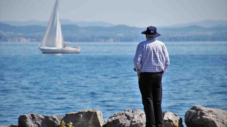 A man wearing a hat watches from shore as a sailboat passes by.