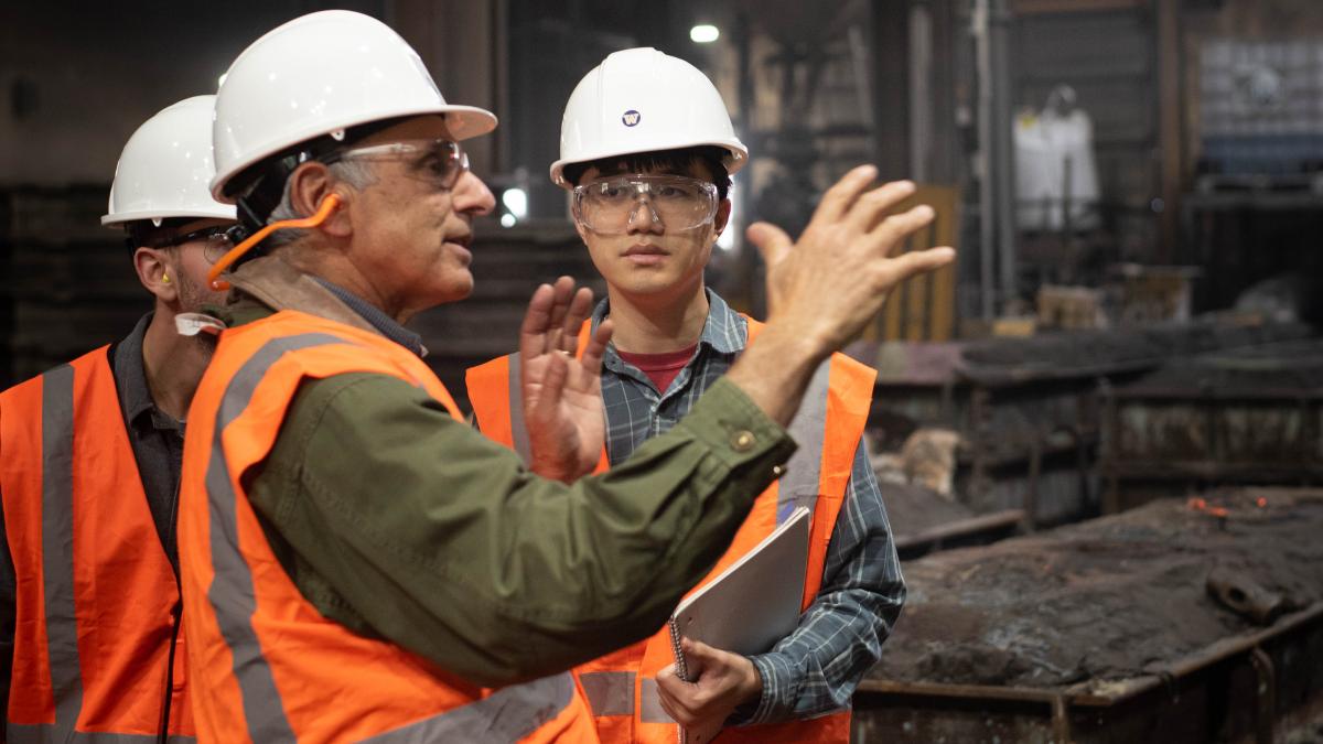 3 men in orange safety vests and hard hats stand in a manufacturing facility.