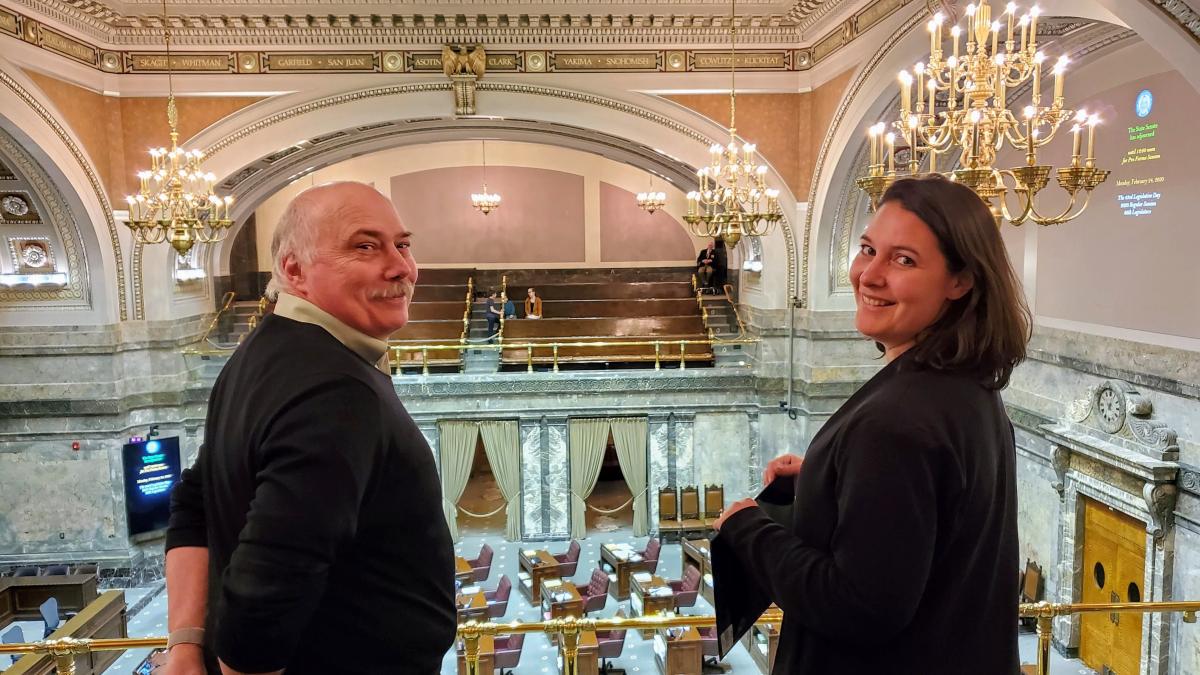 A man and a woman stand in an ornate gallery above a legislative chamber.