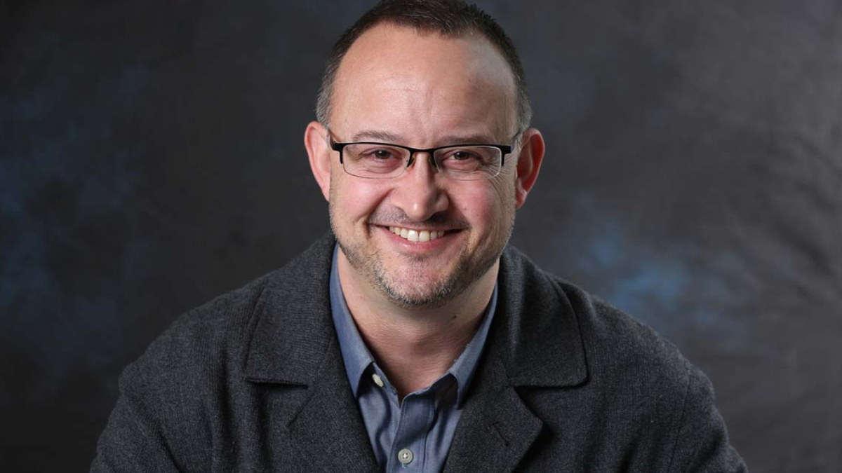 Jeremy Hess smiles at the camera wearing a charcoal gray jacket and blue shirt. He wears glasses and there is a blue/gray backdrop.