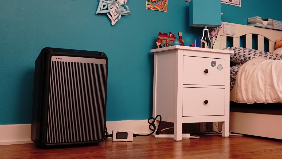 A child's bedroom includes a black air cleaner on the left, a white nightstand and a bed. Teal walls contain many colorful decorations.