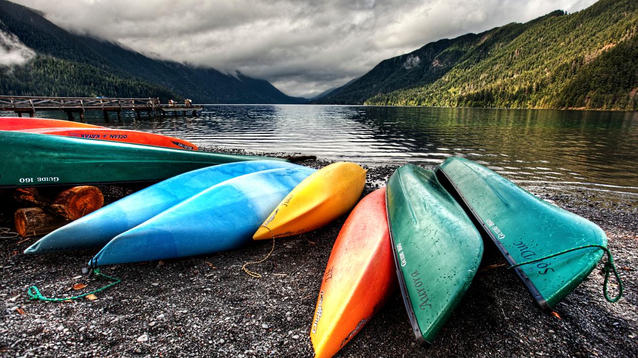 Red, green, yellow and blue canoes on the shore of a lake with green mountains in the background.