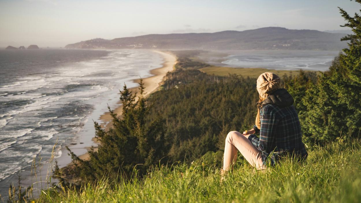 A person sits on a bluff looking out at the ocean and headlands in the distance.