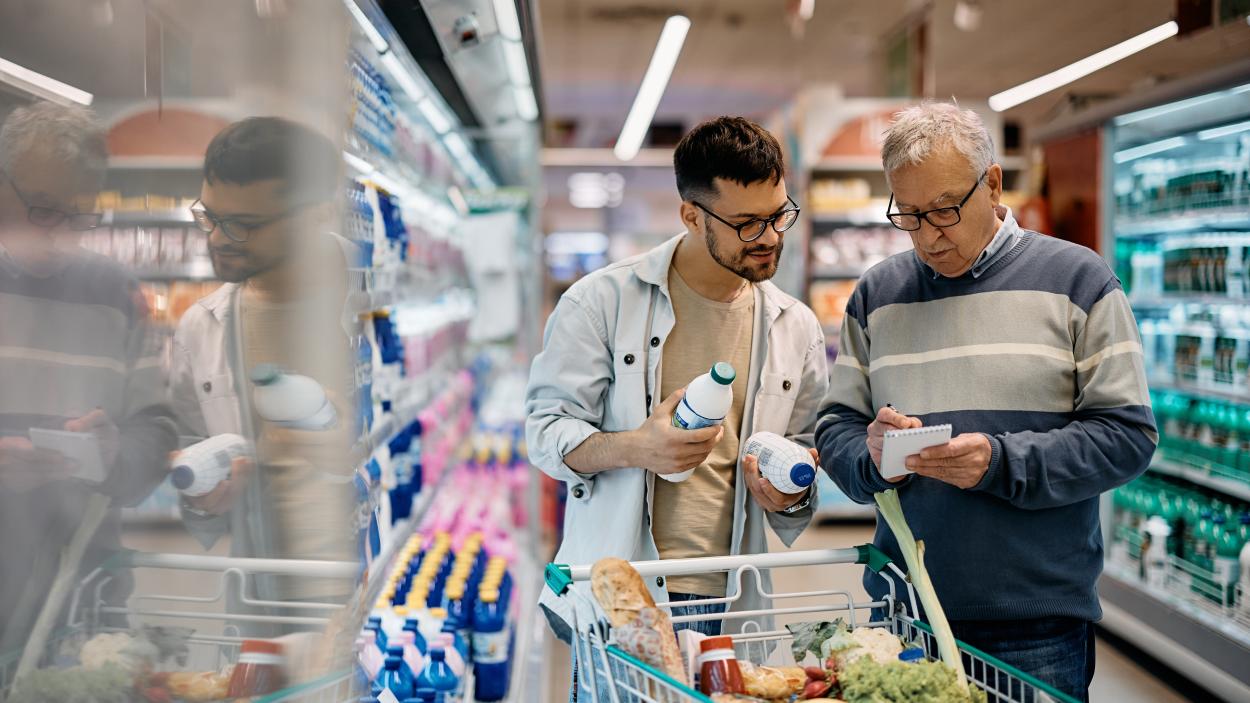 A younger man and an older man shop together in a grocery store, with the older man holding a grocery list on a pad and the young man holding two small milk bottles.