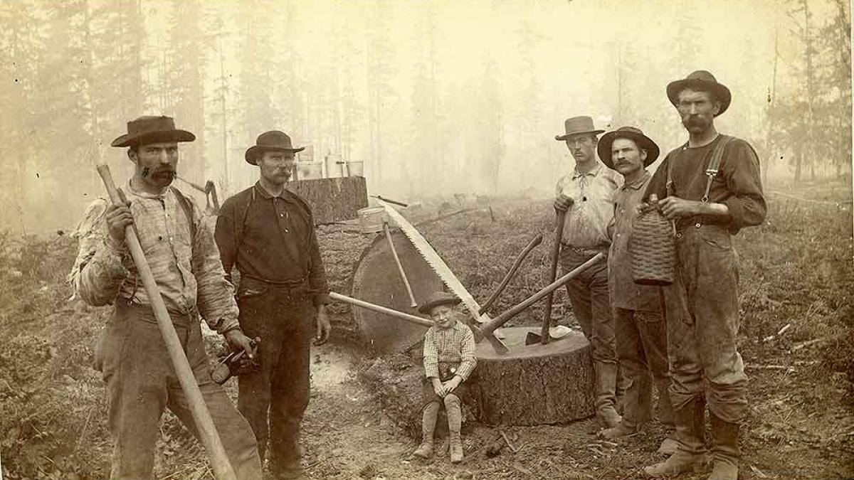 Black and white image of five loggers and a boy in Western Washington, W. T. The men are standing with axes in hand appearing to have just cut down a tree. The boy sits on the stump.