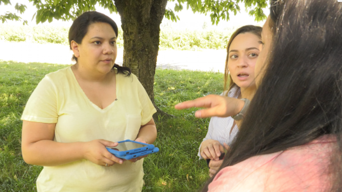 Three women stand outside talking, one holding a computer tablet. 