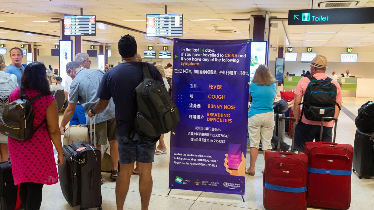 A group of travelers with suitcases stand in line at an airport near a sign about the novel coronavirus
