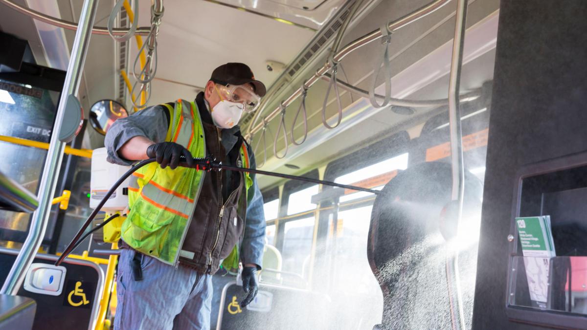 A man in a face mask and protective gear uses a spray wand to spray disinfectant inside a bus.