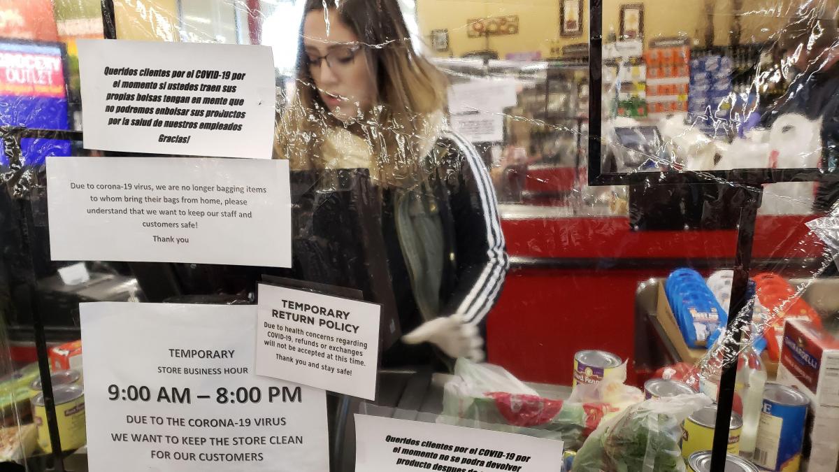 A grocery store clerk stands behind a clear plastic shield filled with signs about COVID-19 while scanning groceries..
