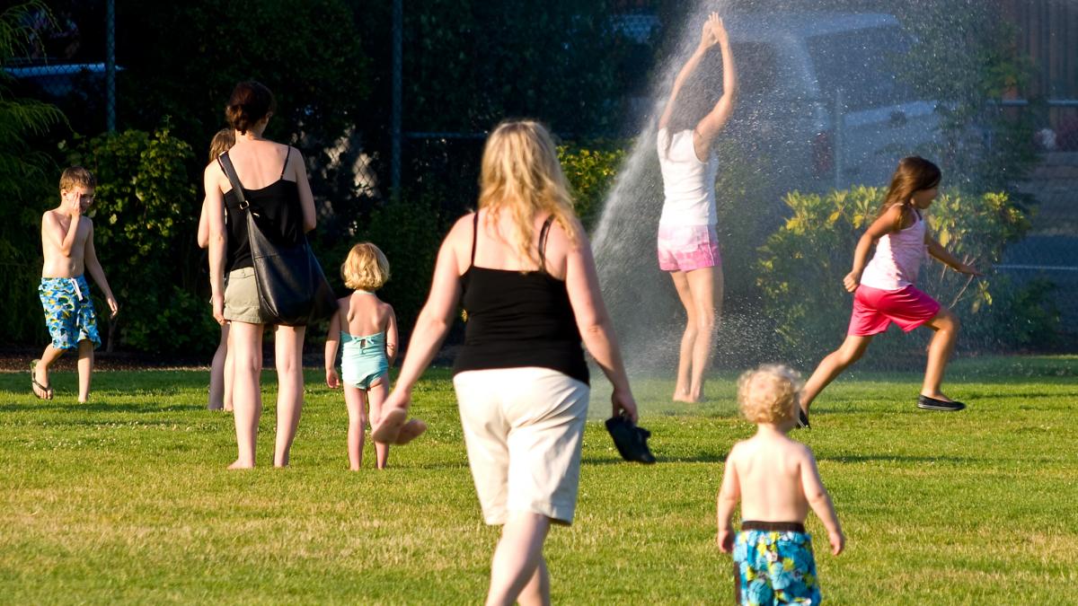 Kids cool off in a sprinkler on a hot day at a park as adults look on.