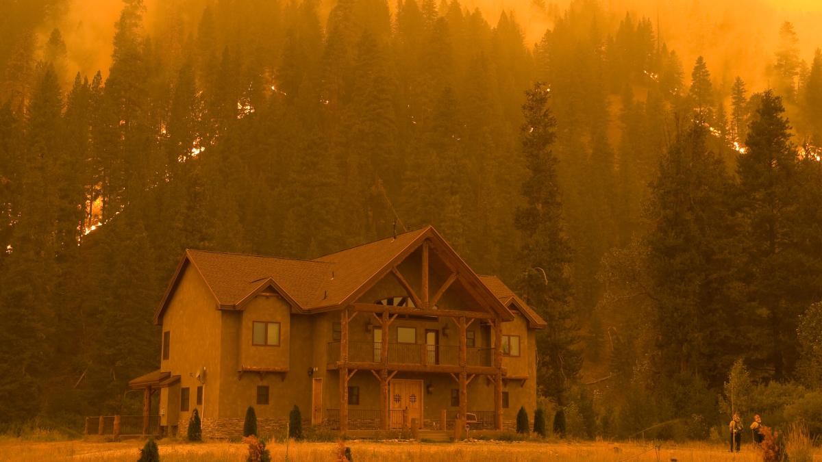 A large home sits in the foreground with wildfire and smoke on the hillside behind it.