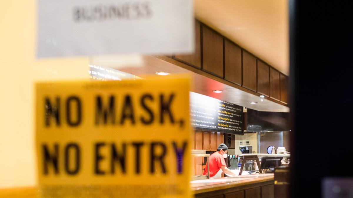 A sign on a door reads "No Mask No Entry" as a worker in a face mask cleans a counter in the background.