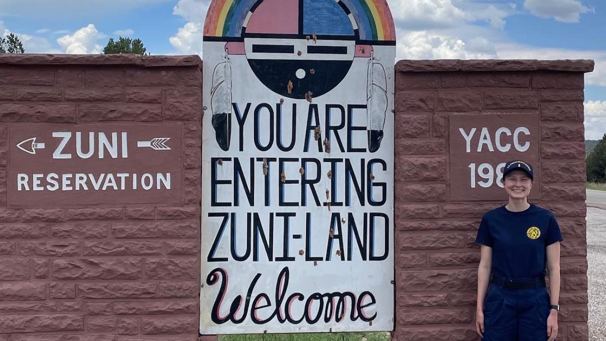 Greta Gunning stands smiling in a JRCOSTEP uniform in front of a sign reading "Zuni Reservation. You are Entering Zuni-Land. Welcome"
