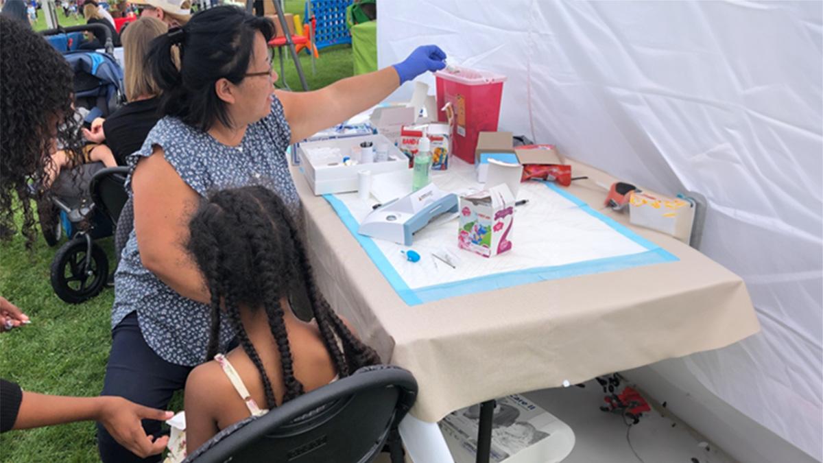 A child sits with a nurse and caregiver after getting a finger prick blood test at a lead testing event.