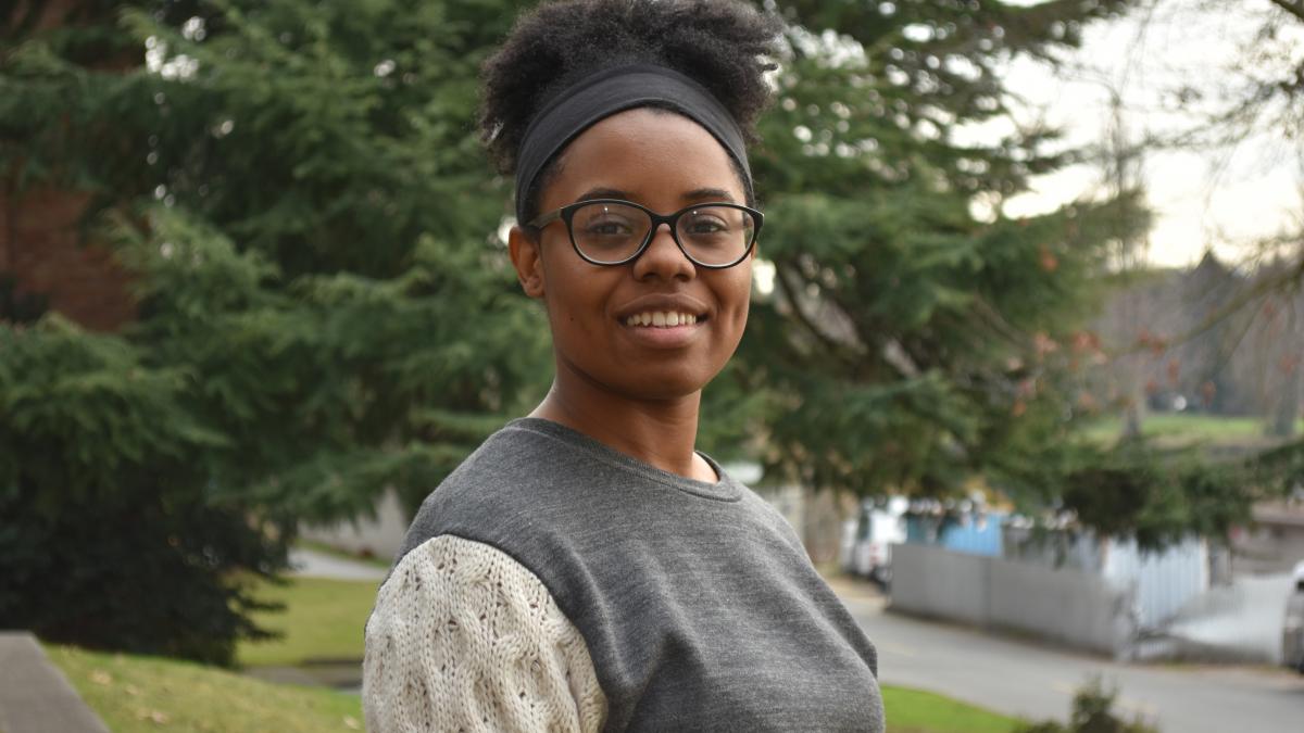 A young woman wearing a gray sweater smiles against a backdrop of trees.