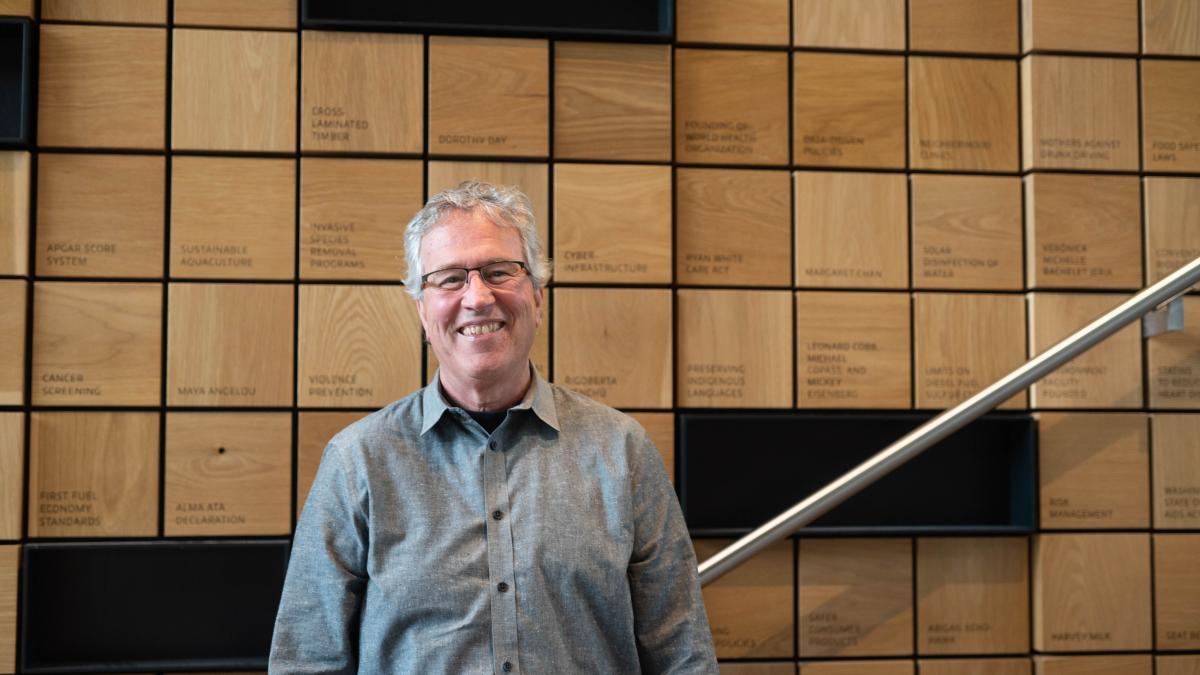 Thomas Burbacher stands smiling in front of a wall of wood blocks inscribed with public health concepts.