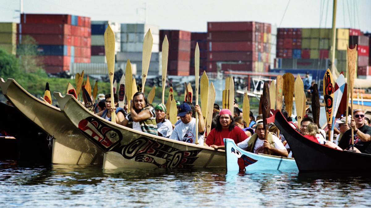A group of people paddle Native boats on a river past a shipping terminal.
