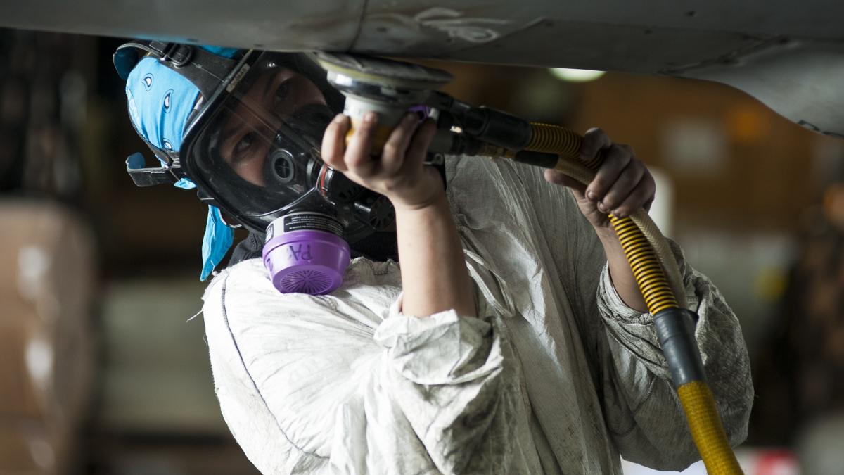 A woman wearing safety gear and an air mask grinds metal fuselage.