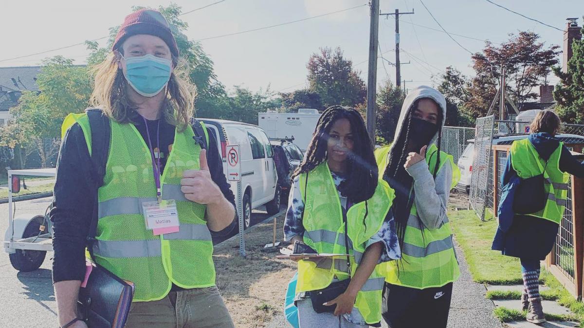 Three youth volunteers from Duwamish Valley Youth Corps smile wearing yellow safety vests, two giving a thumbs up, in a Duwamish Valley neighborhood in Seattle.