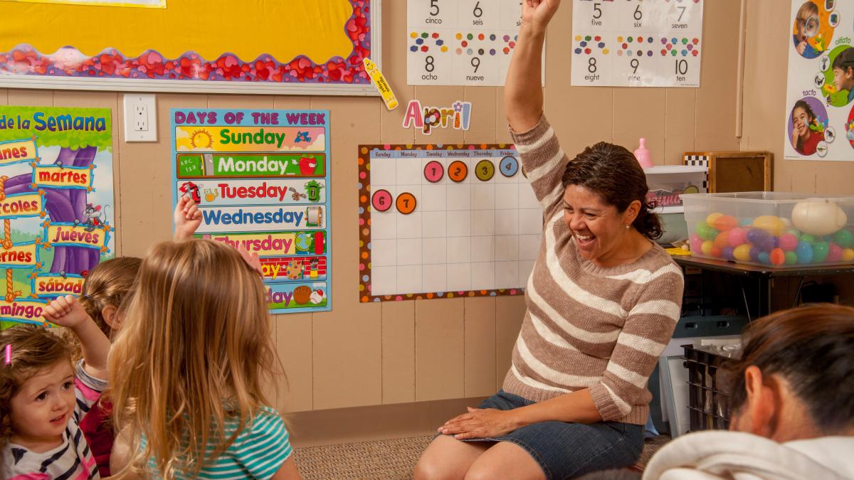 A child care provider sits on the floor raising her hand and smiling with a circle of preschoolers watching.