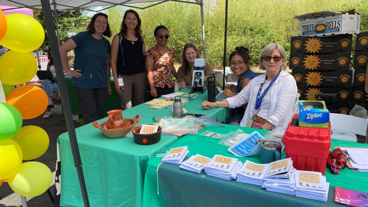 Maegan Chua sits at a table with others, smiling at the camera. On the table are fliers, a clip board, water bottles, a decorative pot, a red plastic tool box and a box of ziploc bags. Yellow, orange and green balloons are verically along the left side of the photo. Boxes are stacked in the background. It is clear those in the photo are at a table, participating in a fair or festival, handing out information. Green tablecloths are on the tables.