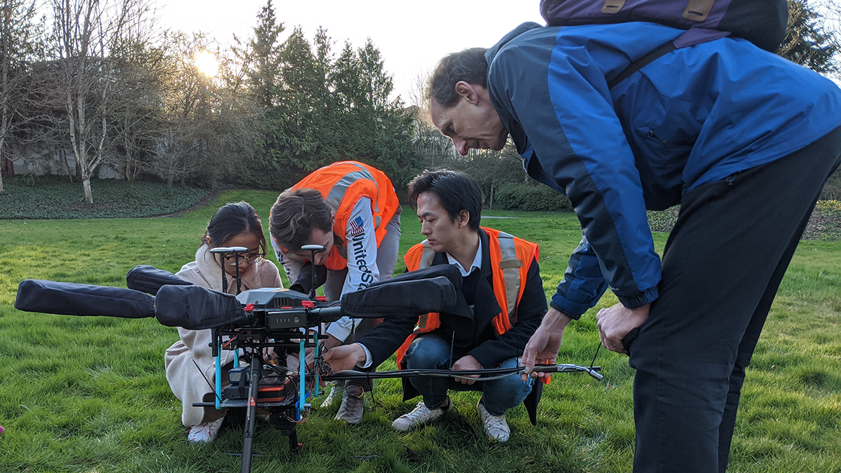Four people crouching around a large drone in a field surrounded by trees.