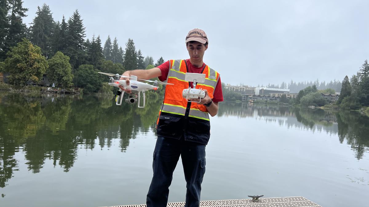 Joey Teresi stands on a dock holding a drone and remote control with a lake in the background.