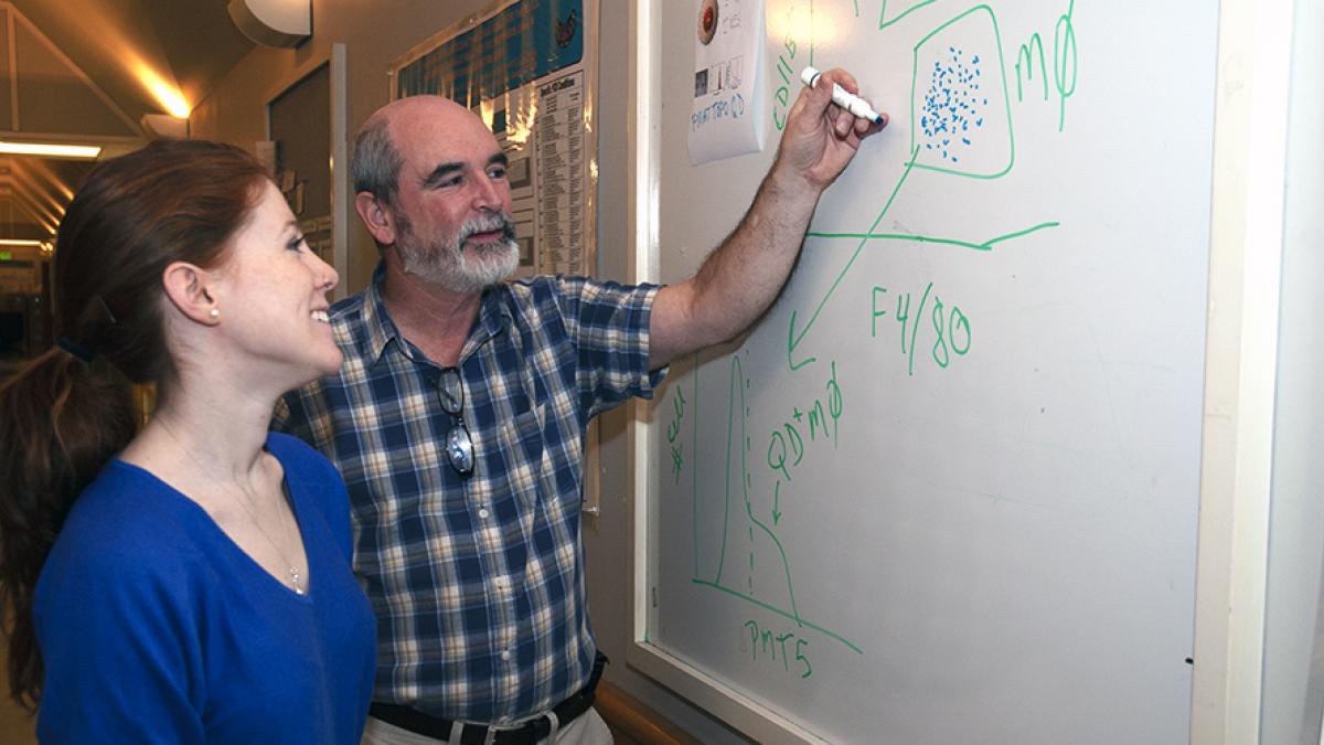Professor Terrance Kavanagh writes on a whiteboard as a student watches.