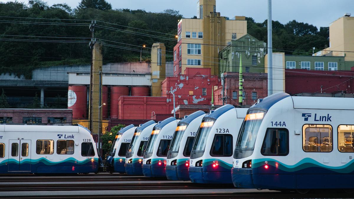 Link light rail cars lined up at a maintenance facility with colorful buildings behind them.