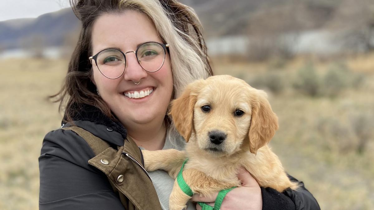 Woman with glasses and a nose ring holds a golden retriever puppy outside while smiling.